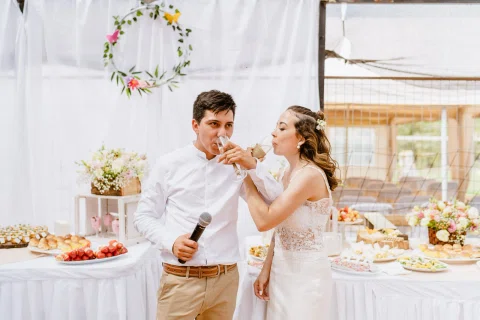 Hombre con camisa blanca y mujer con vestido blanco beben de sus copas al mismo tiempo; él sostiene un micrófono frente a una mesa con comida.