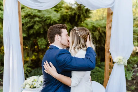 Hombre con traje azul y mujer vestida de blanco se besan bajo una estructura con telas blancas.