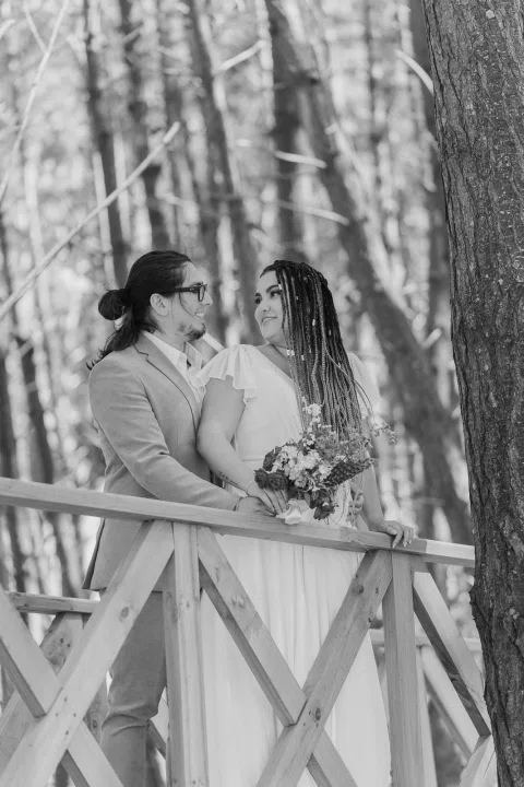 Hombre y mujer se besan junto a una baranda de madera entre árboles, en blanco y negro.