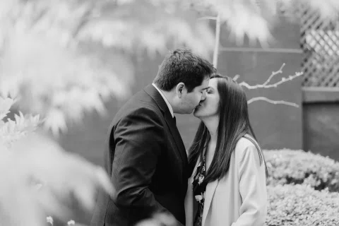 Hombre con traje oscuro y mujer de chaqueta clara se besan en un jardín en una fotografía en blanco y negro.