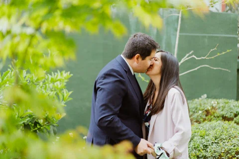 Hombre con traje oscuro y mujer de chaqueta clara se besan en un jardín entre hojas verdes en primer plano.