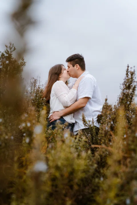 Hombre y mujer se besan entre vegetación alta en un retrato vertical.