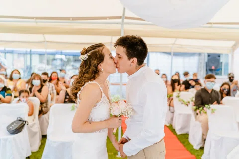 Hombre y mujer se besan bajo una carpa, con varias personas sentadas al fondo.