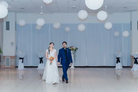 Hombre con traje azul y mujer con vestido blanco caminan juntos por el centro de un salón amplio.