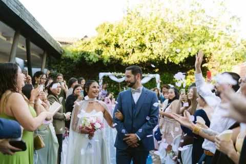 Hombre con traje azul y mujer con vestido blanco caminan entre personas que levantan las manos.