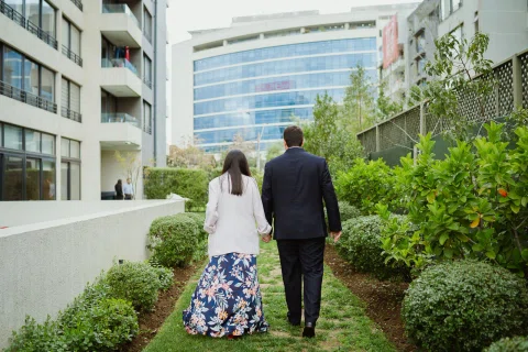 Hombre con traje oscuro y mujer de chaqueta clara caminan de espaldas por un sendero de jardín entre edificios.