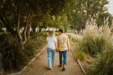 Hombre y mujer caminan de espaldas tomados de la mano por un sendero entre árboles y pastos altos.