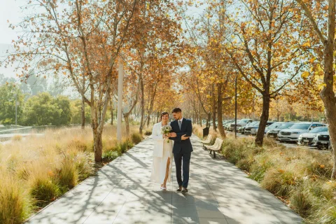 Hombre con traje azul y mujer con traje claro caminan juntos hacia la cámara por un sendero arbolado.