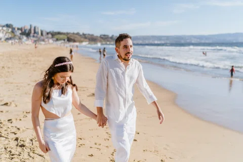 Hombre y mujer caminan tomados de la mano por la playa junto a la línea del agua.