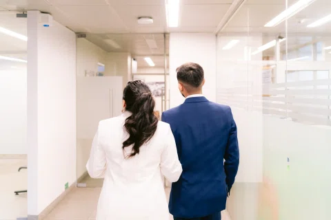 Hombre con traje azul y mujer con traje claro caminan de espaldas por un pasillo blanco.