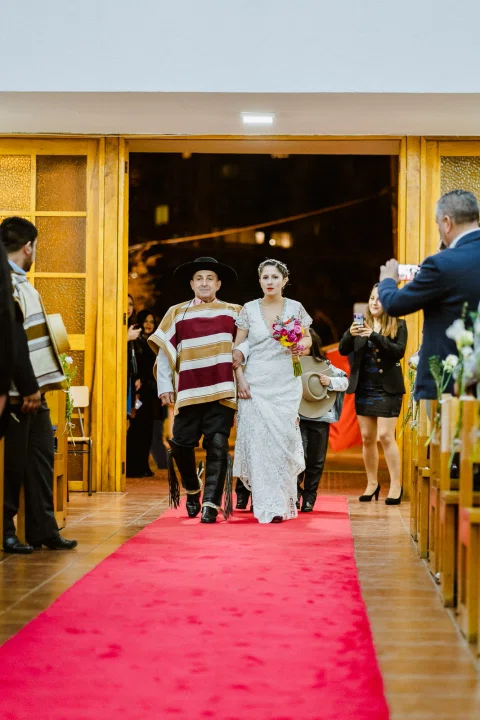 Hombre con poncho y sombrero y mujer con vestido blanco caminan por un pasillo rojo dentro de una iglesia, con otras personas detrás.