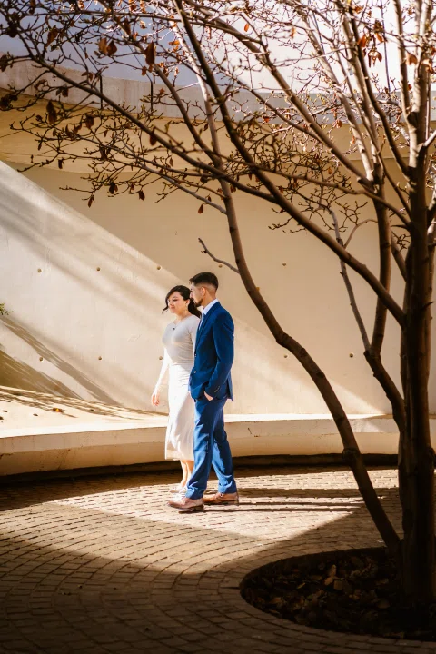 Hombre con traje azul y mujer con traje claro caminan juntos por un patio curvo junto a un árbol.