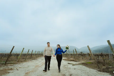Hombre y mujer caminan tomados de la mano por un camino entre viñas; ella sostiene un ramo de flores.