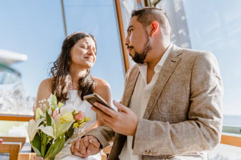 Hombre y mujer conversan junto a un ventanal mientras él sostiene un teléfono y ella un ramo.