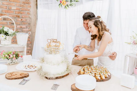 Hombre con camisa blanca y mujer con vestido blanco cortan juntos una torta blanca sobre una mesa con postres y flores.