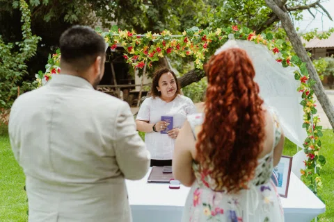 Hombre con chaqueta clara y mujer de cabello rojo permanecen de espaldas frente a una mujer con vestido blanco bajo un arco floral.