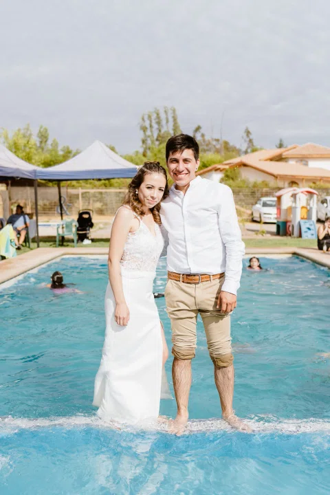 Hombre con camisa blanca y mujer con vestido blanco permanecen de pie dentro de una piscina, con el agua a la altura de las piernas.