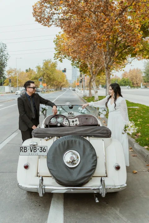 Hombre y mujer permanecen de pie junto a un automóvil convertible blanco visto desde atrás.