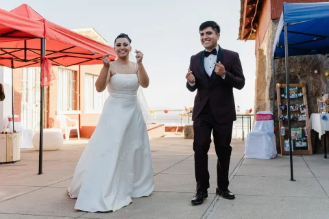 Hombre y mujer están de pie en un patio exterior y levantan una mano mientras sonríen.