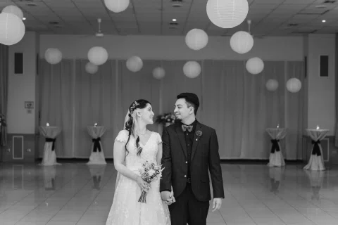 Hombre con traje oscuro y mujer con vestido blanco permanecen de pie tomados de la mano y se miran, en una fotografía en blanco y negro.