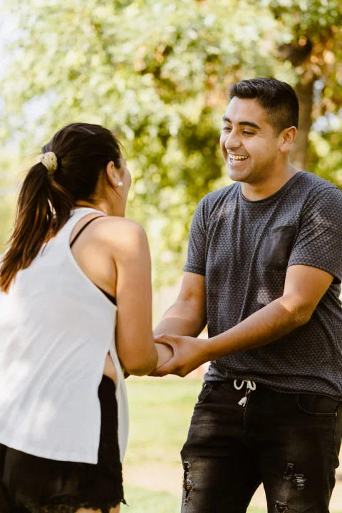 Hombre y mujer permanecen de pie mirándose y tomados de las manos en un parque.