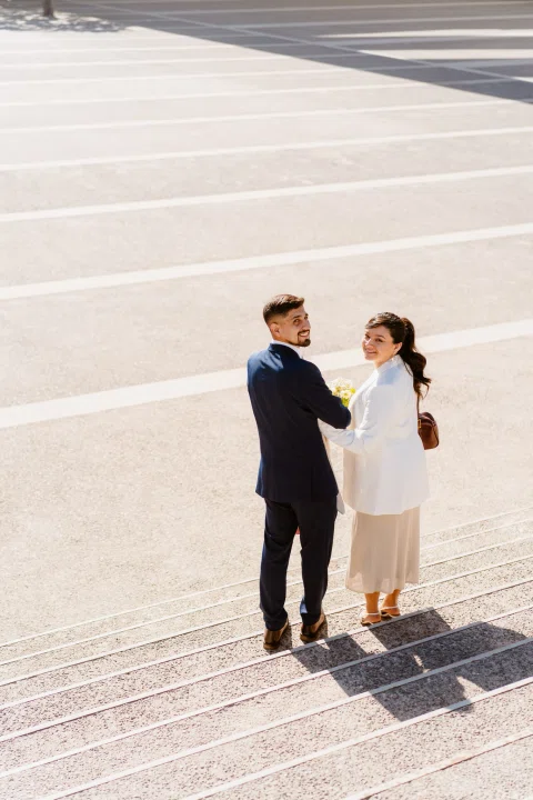 Hombre con traje azul y mujer con traje claro aparecen juntos a distancia sobre una amplia superficie pavimentada.