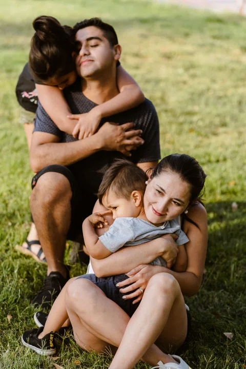 Hombre y mujer están sentados sobre el césped; una niña abraza al hombre desde atrás y la mujer sostiene a un niño pequeño.
