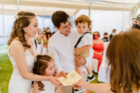 Hombre con camisa blanca sostiene a un niño pequeño mientras una mujer con vestido blanco y una niña reciben una libreta de otra persona.