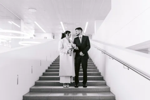 Hombre con traje oscuro y mujer con traje claro posan en una escalera interior, en una fotografía en blanco y negro.