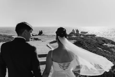 Hombre y mujer están de espaldas frente a la costa mientras el velo de ella se mueve con el viento, en blanco y negro.