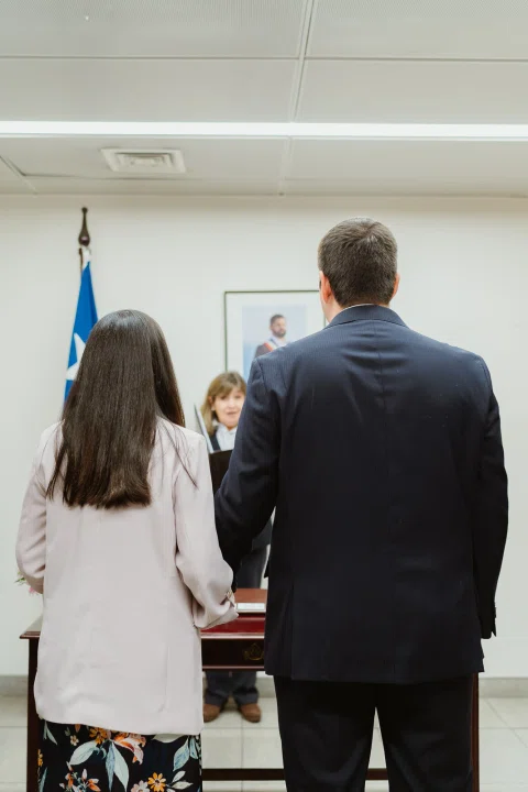 Vista vertical de un hombre con traje oscuro y una mujer de chaqueta clara de espaldas frente a una mesa y una bandera de Chile.