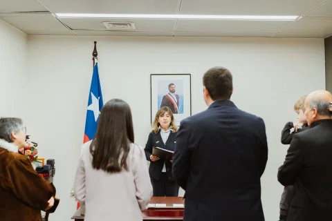 Hombre con traje oscuro y mujer de chaqueta clara son vistos de espaldas frente a una mujer situada detrás de una mesa, con una bandera de Chile al costado.