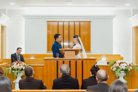Hombre con traje azul y mujer con vestido blanco permanecen frente a frente junto a un atril, con personas sentadas delante.