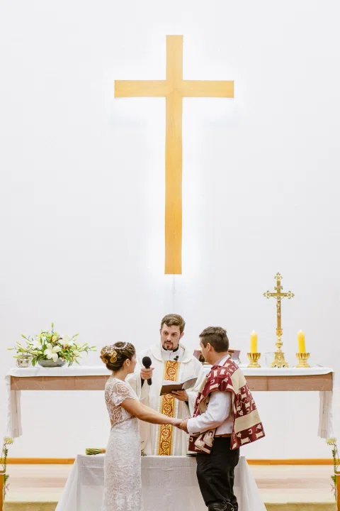 Hombre con poncho y mujer con vestido blanco están frente al altar bajo una gran cruz dorada.