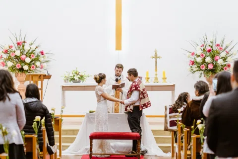 Hombre con poncho y mujer con vestido blanco están de pie frente al altar; detrás se ve un hombre con vestimenta litúrgica.