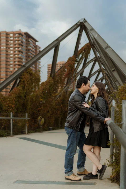 Hombre y mujer permanecen frente a frente muy próximos junto a la baranda de un puente metálico.