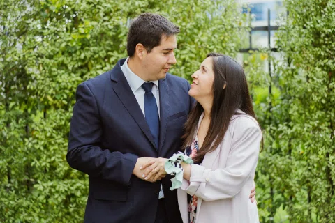 Hombre con traje oscuro y mujer de chaqueta clara están frente a frente en un jardín; ella sostiene un pequeño ramo.