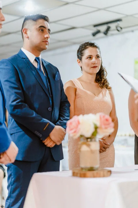Hombre y mujer permanecen de pie frente a una mesa con flores en un espacio interior, con otras personas alrededor.