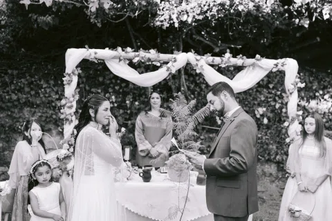 Hombre con traje oscuro y mujer con vestido blanco permanecen frente a una mujer con vestido morado bajo un arco floral, en blanco y negro.