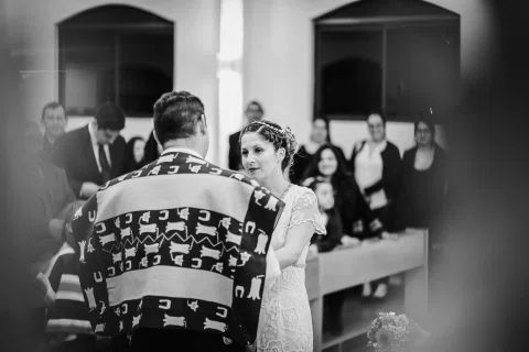 Mujer con vestido blanco está frente a un hombre con poncho, con varias personas detrás, en una fotografía en blanco y negro.