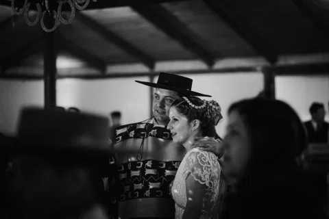 Hombre con poncho y sombrero mantiene un brazo alrededor de una mujer con vestido blanco, en una fotografía en blanco y negro dentro de un salón.