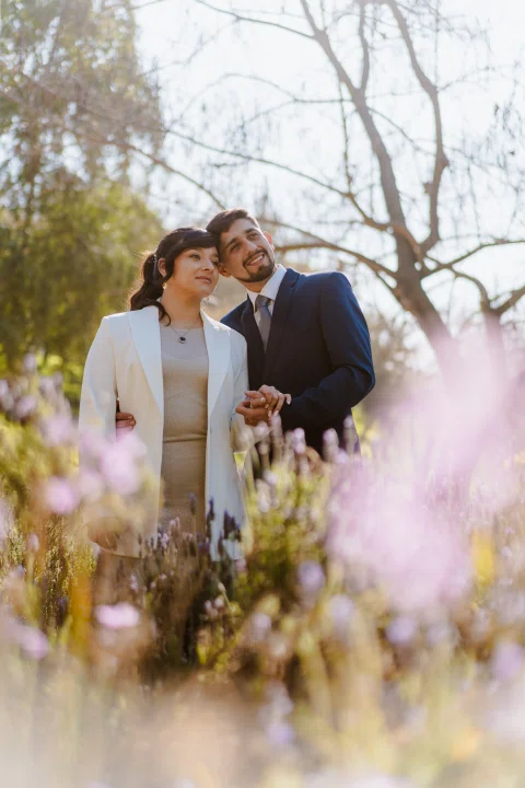 Hombre con traje azul y mujer con traje claro permanecen juntos entre flores y vegetación del parque.