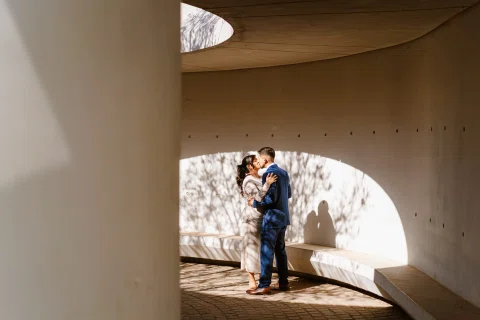 Hombre con traje azul y mujer con traje claro permanecen juntos dentro de una estructura curva marcada por luces y sombras.