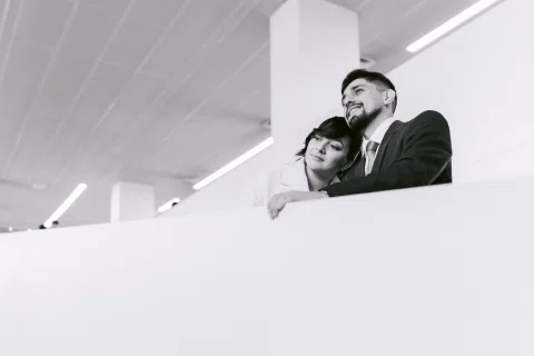 Hombre con traje oscuro y mujer con traje claro permanecen juntos junto a un muro blanco, en una fotografía en blanco y negro.
