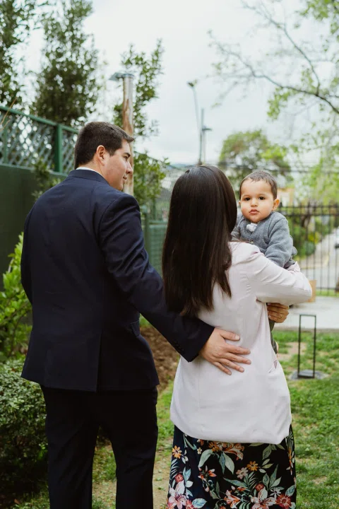 Hombre con traje oscuro y mujer de chaqueta clara están juntos en un jardín mientras ella sostiene a un niño pequeño en brazos.