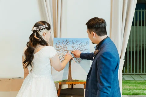 Hombre con traje azul y mujer con vestido blanco marcan con sus dedos un corazón dibujado en el tronco de un árbol pintado sobre un lienzo.