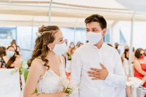 Hombre y mujer con mascarillas permanecen de pie uno junto al otro bajo una carpa, con varias personas sentadas detrás.