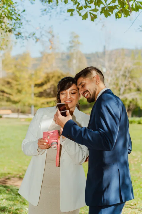Hombre con traje azul y mujer con traje claro miran juntos una fotografía instantánea mientras ella sostiene la cámara rosada.