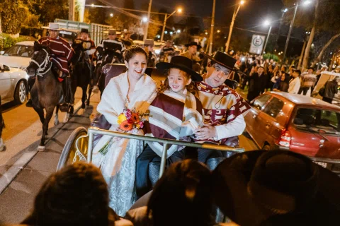 Mujer con vestido blanco, niño con poncho y sombrero y hombre con poncho y sombrero viajan sentados en un carruaje tirado por caballos durante la noche.