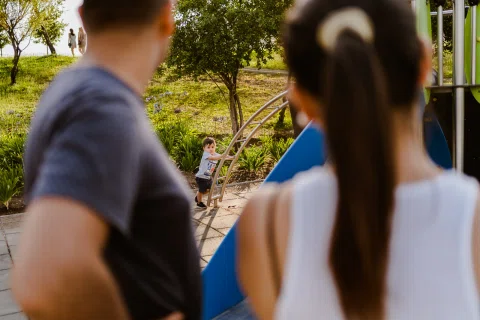 Hombre y mujer vistos de espaldas observan a un niño junto a una estructura de juegos.